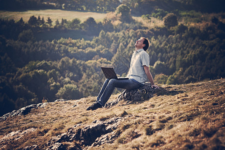 年轻人用笔记本电脑坐在山坡的草地上地平线沉思男性商业休息寂寞微笑石头技术天空图片