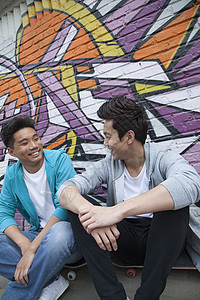 两个年轻人坐在他们的滑板上 挂在墙前涂着涂鸦的男孩子身上团结棕色服装青年乐趣短发活力幸福文化城市背景图片