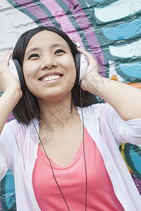 年轻微笑的女士拿着她的耳机 同时享受着在墙壁前用涂鸦听音乐城市听力头发城市生活休闲收腰活力摄影服装快乐图片