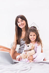 带笔记本电脑的幸福母亲和女儿玩具熊孩子技术卧室玩具睡衣女孩羽绒被公寓女士图片