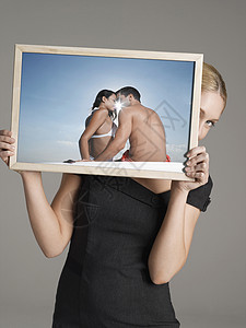 年轻女商务人士在海滩上用灰色背景偷看情侣照片后边的相片图片
