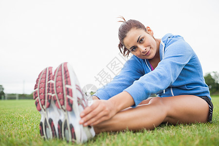 漂亮的运动型女子在坐在草地上时伸展双腿双手棕色训练女士调子女性热身夹克运动服头发图片
