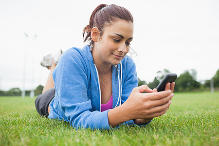 拥有手机的漂亮女人躺在草地上电话夹克蓝色短信公园微笑阳光棕色女士天空图片