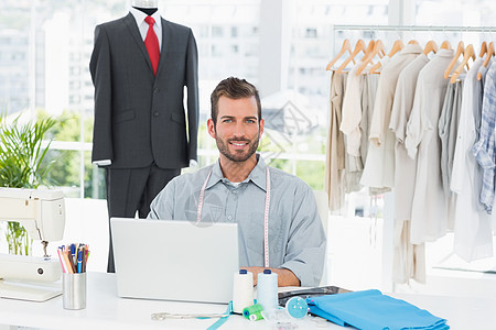 在工作室使用笔记本电脑的男性时装设计员微笑裁缝织物工作造型师配件男人卷尺设计师技术衣架图片