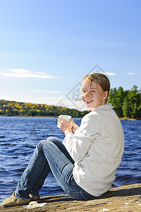 年轻女子在湖岸休闲晴天杯子岩石荒野青年风景享受蓝色饮料女士图片