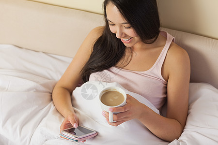 坐在床上用智能手机发短信和喝咖啡的年轻女士 她们欢欣鼓舞地坐在床上图片
