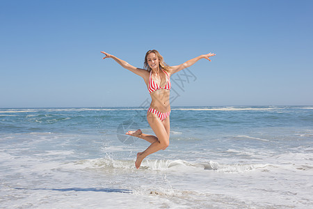 穿着比基尼的漂亮美女在海滩上跳跃浅色条纹头发支撑自由女士沙滩海岸线微笑乐趣图片