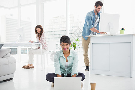 使用笔记本电脑坐在地板上微笑的女商务人士工作团队快乐混血杯子桌子办公室男性女性热饮图片