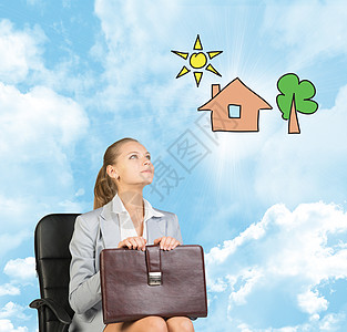 穿着裙子 衬衫和夹克的女商务人士坐在椅子上 拿着公文包想象着有树的房子 以蓝天 白云为背景图片