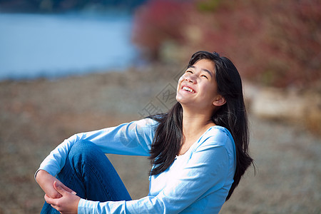 穿着蓝衬衫和牛仔裤的少女女孩坐在岩石湖边长发混血青少年支撑黑发混血儿女性牛仔布靴子巨石图片