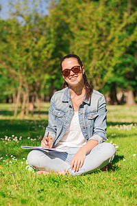 带着笔记本写作在公园的年轻微笑女孩学习工作簿快乐铅笔女士牛仔布教育头发太阳镜幸福图片