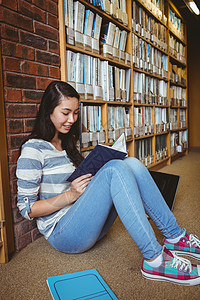 坐在地板上坐在图书馆阅读书墙壁对面的微笑学生文学女士休闲学校高等教育大学生校园知识混血快乐图片