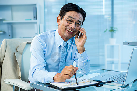 笑笑的亚洲商务人士给某人打电话沟通写作人士旋转办公室电脑电话夹克手机专注图片