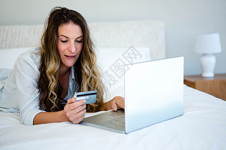妇女躺在她床上的笔记本电脑上金发女郎喜悦网上购物羽绒被享受快乐技术娱乐性坐垫电子商务图片