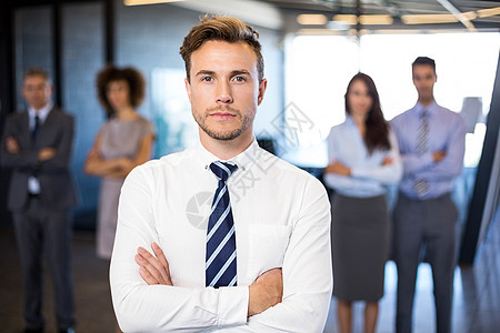 商务人士对着镜头微笑 而他的同事则站在背景上站着职场管理人员伙伴团队领导男性办公室成功领导者混血图片