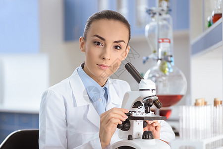 年轻女科学家穿着实验室外衣 与显微镜一起工作 并观看照相机图片