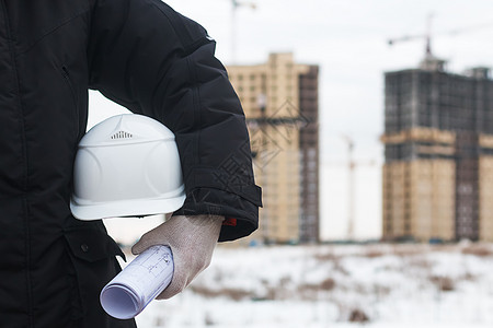 建筑师或工程师在新的高层公寓楼和建筑起重机的背景下为工人安全手持黄色头盔在冬季景观背景下剪影起重机举起负载图片