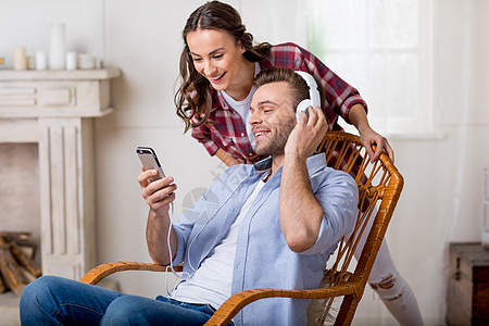 男人在耳机里笑着微笑 在智能手机上展示一些东西给后面的快乐女人女朋友男朋友摇椅女士椅子夫妻手势友谊成人水平图片