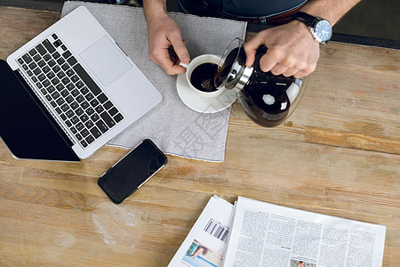 男人用笔记本电脑和报纸把咖啡倒在桌子上的杯子里的部分观点裁剪早餐手机商业自由职业者成人职业饮料商务远程图片