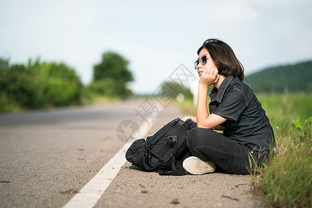 妇女坐在农村一条公路上搭便车的背包车上短发女孩女士青少年远足太阳镜天空旅游路线游客图片