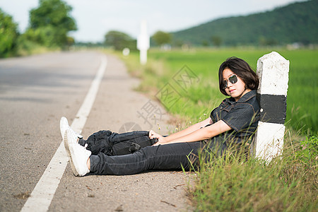 妇女坐在农村一条公路上搭便车的背包车上游客背包太阳镜女性冒险短发女士假期牛仔裤女孩图片