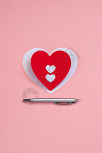 粉红色背景上带银笔的红纸心 情人节或婚礼日明信片概念图片