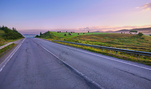 宽阔的沥青道路 边边缘有压土机 在绿平原之间行走到山顶 远在地平线上图片