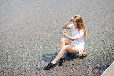 坐在路边的滑板上时 年轻美丽的笑笑女孩用智能手机做自拍运动木板金发女郎溜冰者牛仔裤沥青城市滑冰短裤青少年图片