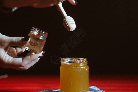 玻璃罐中的蜂蜜紧靠在红色木桌上的黑色背景上 用棒状蜂蜜勺倒蜂蜜桌子橙子产品花蜜营养养蜂业作品乡村生物杏仁图片