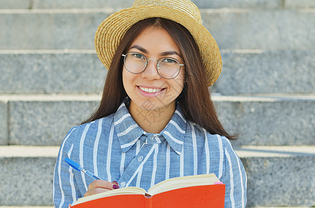 一名戴眼镜的亚洲青年学生身穿蓝条纹衬衫戴顶帽子的肖像青少年喜悦电脑女孩牛仔裤女性快乐楼梯城市大学图片