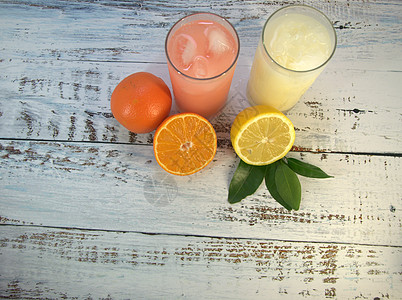 两杯玻璃杯 上面放着清爽的果汁和冰块 放在一个纺织品支架上 把一半的橙子和叶子切成两半 在一张白色的木桌上放着一半的柠檬汁娱乐假图片