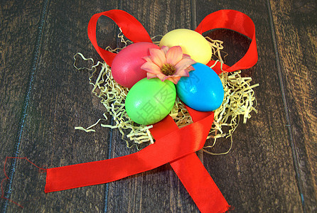 四个漆成复活节的鸡蛋在稻草巢中 中间有一朵花 躺在木制桌子上的红色丝带上传统装饰丝带蓝色风格丝绸海报手工庆典食物图片