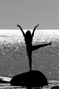 一个女孩站在海中的一块石头上 双臂和双腿举起的剪影 在海面上波光粼粼的阳光下图片