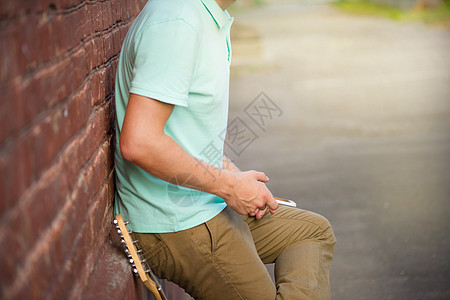 夏天 派对 年轻时尚的家伙 站在棕砖墙附近 手拿着手机 在吉他旁边 阳光运动鞋期望男生男士短信电话家具衬衫裤子检查图片