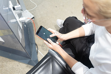 一名年轻女子坐在充电站前 看着她的智能手机 在机场的免费充电站为手机充电力量民众插头充电器电缆休息室技术适配器桌子大厅图片