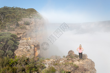 女徒步者站在岩石顶峰上 山谷中浓雾升起图片