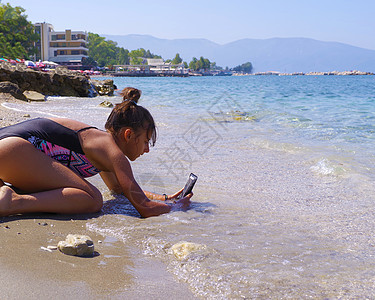 年轻漂亮的女孩坐在海边的海滩上 用手机拍照 夏季 大海和沙滩图片