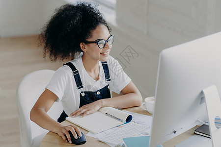 高兴的年轻非洲裔美国女性坐在现代电脑前 做项目工作 桌上有许多文件和记事本 戴着眼镜矫正视力 穿着休闲服图片
