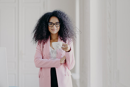 拍摄有非洲发型 喝芳香饮料 戴眼镜 正装 在白色背景下在办公室摆姿势的迷人女性 为您的宣传内容复制空间 人与企业图片