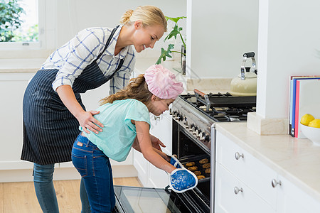 母亲和女儿把饼干盘放在烤箱中快乐围裙公寓手套女士女性厨房服装家庭托盘图片