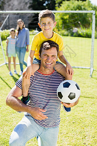 父亲带着足球微笑 他的父亲在公园上肩并肩背着儿子去打橄榄球图片