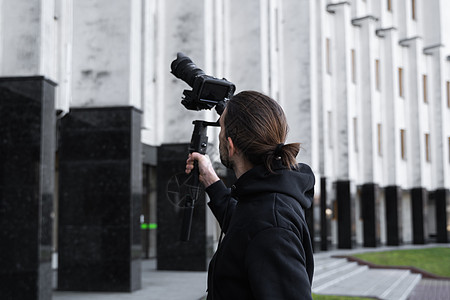 年轻的专业摄像师在 3 轴万向稳定器上手持专业相机 专业设备有助于制作高质量视频而不会抖动 身穿黑色连帽衫的摄影师正在制作视频博图片