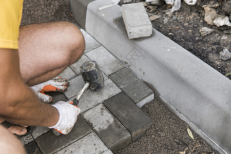 一名建筑工人的手戴着橙色手套 手上拿着锤子配件 正在铺设新的外部铺路石 小心地将一块铺路石放在水平和倾斜的土壤基础上 沙地基 建图片