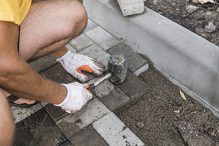 一名建筑工人的手戴着橙色手套 手上拿着锤子配件 正在铺设新的外部铺路石 小心地将一块铺路石放在水平和倾斜的土壤基础上 沙地基 建图片