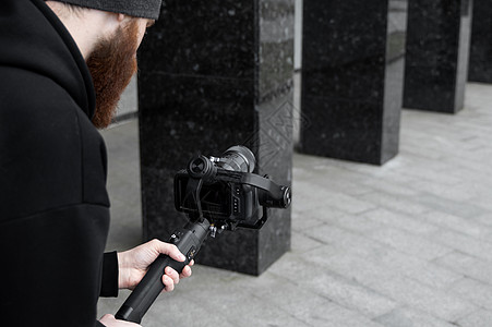身穿黑色连帽衫 留着胡子的专业摄像师 手持专业相机 安装在 3 轴万向稳定器上 电影制作人使用专业的电影摄影机制作精彩视频 电影图片