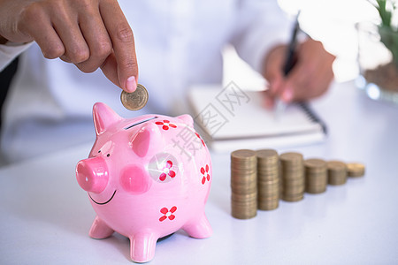 商务人士的手将硬币放入粉红色的存钱罐中 手正要写一个收入和支出帐户 财务会计概念 计算业务成功的资产图片