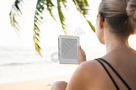 女性在海边户外拥有电子阅读器 其背景是棕榈树 在旅行中放松并享受着在户外阅读最喜欢的书籍闲暇触摸屏读者药片假期成人日落酒店女士女图片