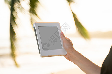 女性在海边户外拥有电子阅读器 其背景是棕榈树 在旅行中放松并享受着在户外阅读最喜欢的书籍青少年酒店闲暇电脑读者女孩日落触摸屏屏幕图片
