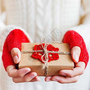 穿着白色针织毛衣和手套的女人手里拿着一份用牛皮纸包装的礼物 上面有钩编的红色雪花 DIY 圣诞节或新年 情人节礼物图片