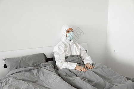 身穿保护性白色西装和面罩的人的肖像 他们因为2019年风湿而坐在卧室的床上 等待冠状病毒检疫结束 自我隔离感染人士安全危险男人防图片